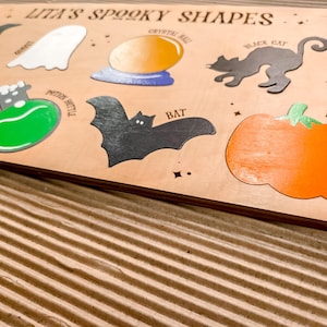 May include: Wooden puzzle board with the text "Lita's Spooky Shapes". Features cut-out shapes including a witch's hat, ghost, crystal ball, black cat, potion bottle, bat, and pumpkin. The board is light brown with colourful accents.