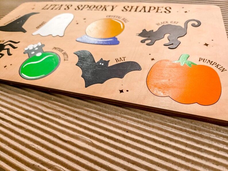 May include: Wooden puzzle board with the text "Lita's Spooky Shapes". Features cut-out shapes including a witch's hat, ghost, crystal ball, black cat, potion bottle, bat, and pumpkin. The board is light brown with colourful accents.