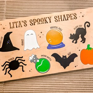 May include: Wooden board with Halloween shapes: witch's hat, ghost, crystal ball, black cat, pumpkin, spider, potion bottle, and bat. The text "Lita's Spooky Shapes" is at the top. The shapes are in black, white, orange, and green.
