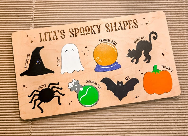 May include: Wooden board with Halloween shapes: witch's hat, ghost, crystal ball, black cat, pumpkin, spider, potion bottle, and bat. The text "Lita's Spooky Shapes" is at the top. The shapes are in black, white, orange, and green.