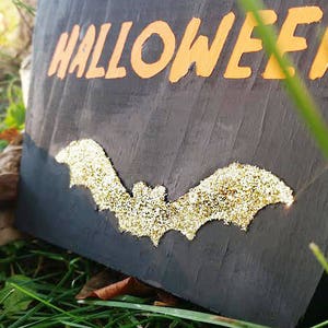 May include: A black wooden Halloween sign with white and orange lettering that reads "Make Yours a Halloween". A gold glitter bat is affixed to the sign. The sign is resting on green grass.