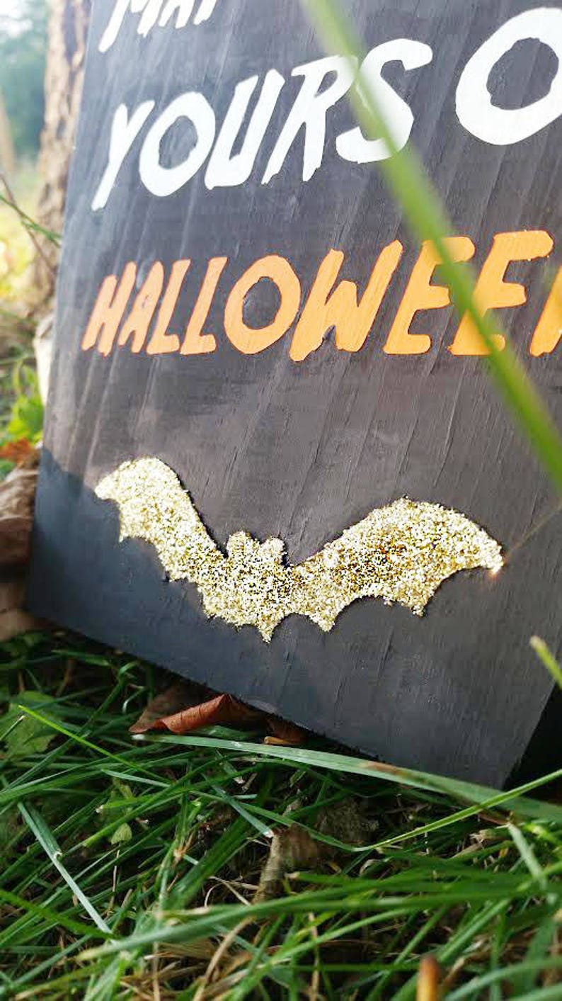May include: A black wooden Halloween sign with white and orange lettering that reads "Make Yours a Halloween". A gold glitter bat is affixed to the sign. The sign is resting on green grass.