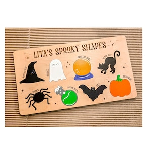 May include: Wooden puzzle board with Halloween shapes: witch's hat, ghost, crystal ball, black cat, spider, potion bottle, bat, and pumpkin. The text "Lita's Spooky Shapes" is printed on the board. The shapes are in black, white, orange, and green.