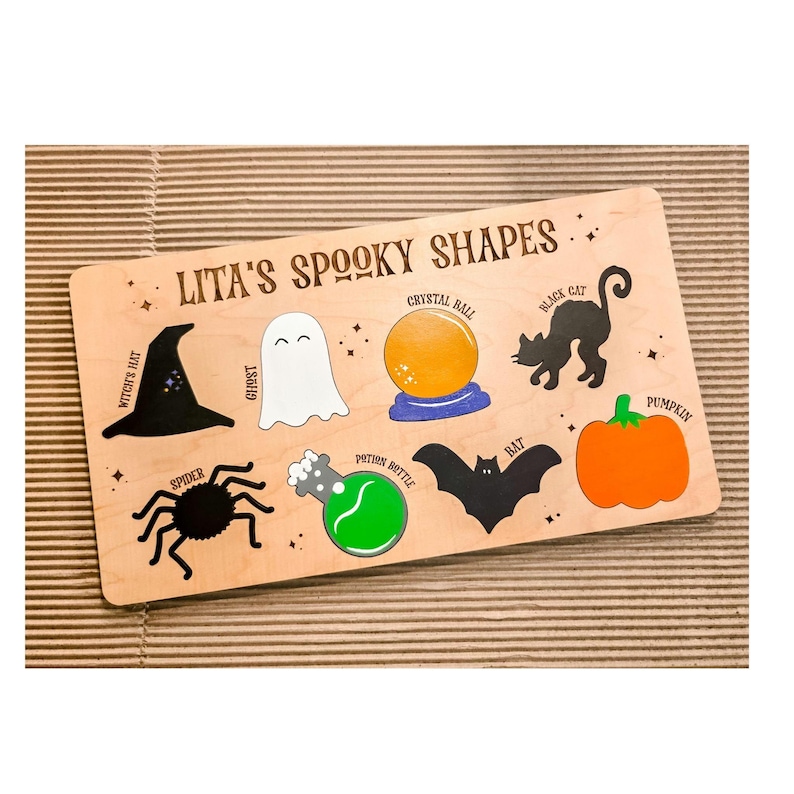 May include: Wooden puzzle board with Halloween shapes: witch's hat, ghost, crystal ball, black cat, spider, potion bottle, bat, and pumpkin. The text "Lita's Spooky Shapes" is printed on the board. The shapes are in black, white, orange, and green.
