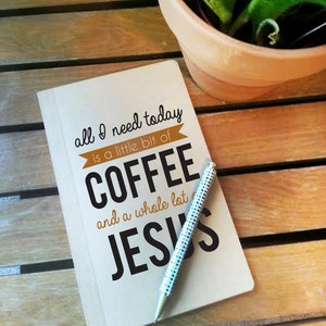 May include: A notebook with the text "all I need today is a little bit of COFFEE and a whole lot of JESUS" and a pen with a white and black polka dot design. The notebook and pen are on a wooden surface, with a potted plant in the background.