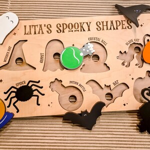May include: Wooden Halloween puzzle featuring cut-out shapes and matching pieces. Includes a ghost, witch's hat, spider, crystal ball, black cat, pumpkin, potion bottle, and bat. The puzzle is labelled "Lita's Spooky Shapes".