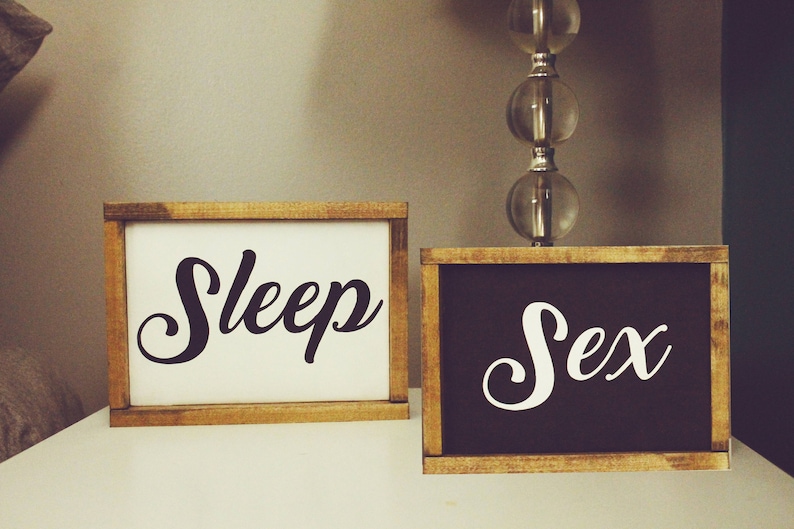 May include: Two framed wooden signs with the words "Sleep" and "Sex" in elegant white script. The "Sleep" sign has a white background, while the "Sex" sign has a black background. The signs are displayed on a white surface.