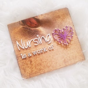 May include: Wooden sign with the text "Nursing is a work of" in white lettering. A purple string art heart is on the right side. The sign is made of stained wood and sits on a white, fluffy surface.