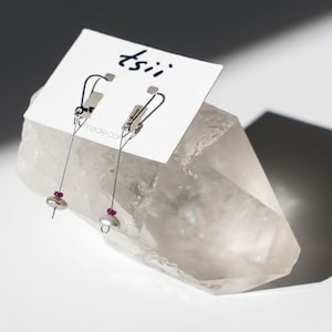 May include: A pair of silver dangle earrings with a small white pearl and a pink gemstone on each earring. The earrings are displayed on a clear crystal.