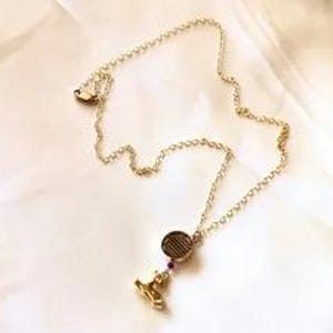 Könnte beinhalten: Eine goldfarbene Kettenhalskette mit einem kleinen, runden Anhänger, der horizontale Linien aufweist. Unter dem runden Anhänger hängt ein kleiner, goldfarbener Charm, der durch eine winzige, dunkelrote Perle akzentuiert wird.