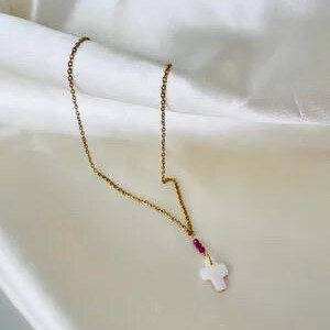 May include: A gold-toned metal chain necklace featuring a small white cross pendant suspended from a single dark pink bead. The delicate link chain connects to the bead, which sits directly above the white cross-shaped charm.