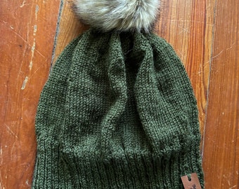May include: A green knitted beanie with a brown leather tag that says "The Greenery Co.". The beanie has a fluffy white pom pom on top.