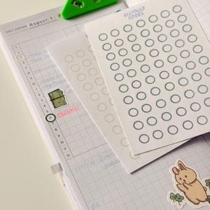 May include: A daily agenda planner with grid paper, a green triangular page marker, and two sticker sheets featuring rows of circles labeled Bestowed Goods. A small sticker of a brown rabbit with green clover leaves is placed on the bottom right corner.