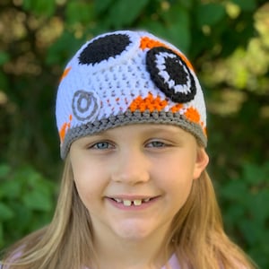 Może przedstawiać: Dziecko w białej, pomarańczowej i szarej czapce na drutach z czarnymi i białymi dzianinowymi kółkami z przodu. Czapka została zaprojektowana tak, aby przypominała postać BB-8 z serii Gwiezdne Wojny.