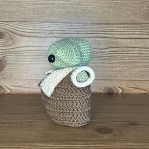 K&ouml;nnte beinhalten: Eine geh&auml;kelte Baby-Yoda-Puppe in Gr&uuml;n und Braun mit einer wei&szlig;en H&auml;keldecke. Die Puppe sitzt auf einer Holzoberfl&auml;che.