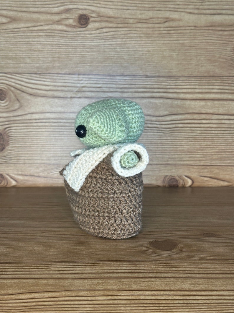 K&ouml;nnte beinhalten: Eine geh&auml;kelte Baby-Yoda-Puppe in Gr&uuml;n und Braun mit einer wei&szlig;en H&auml;keldecke. Die Puppe sitzt auf einer Holzoberfl&auml;che.