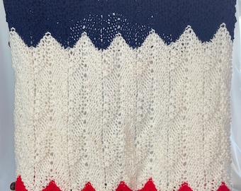 May include: A knitted blanket with a chevron pattern in red, white, and blue colors. The blanket is draped over a chair.