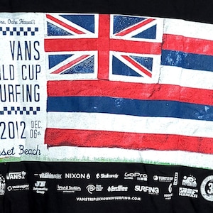 Op de afbeelding: Zwart T-shirt met een ontwerp voor de Vans World Cup of Surfing op Sunset Beach in 2012. Het ontwerp bevat een vlag met rode, witte en blauwe strepen en een Union Jack. Sponsorenlogo's staan onderaan.