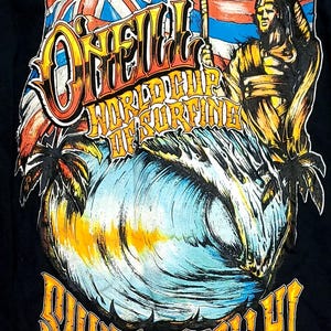 May include: Black t-shirt featuring a colorful graphic of a wave, palm trees, and a surfer with a spear. The design includes the text "O'Neill World Cup of Surfing" and "Sunset Beach HI 2010-2011."