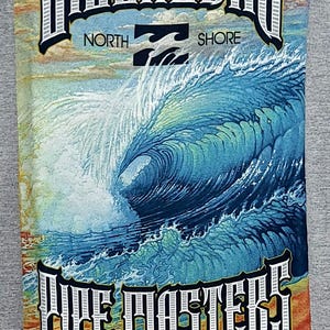 Op de afbeelding: Grijze t-shirt met een levendige afbeelding van een grote oceaangolf en de tekst "BILLABONG NORTH SHORE PIPE MASTERS HAWAII DECEMBER 8-20, 2010". Het ontwerp bevat het Billabong-logo en sponsorlogo's.