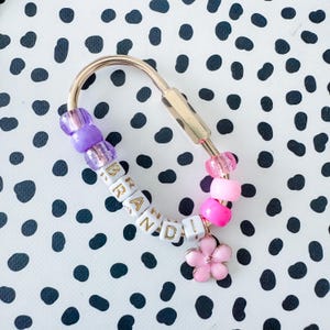 Könnte beinhalten: Ein goldfarbener Schlüsselanhänger mit Perlendesign. Der Schlüsselanhänger trägt den Namen "BRANDI" in weißen Buchstabenperlen. Er enthält lila und rosa Perlen sowie einen rosa Blumenanhänger. Der Hintergrund ist ein schwarz-weißes Tupfenmuster.