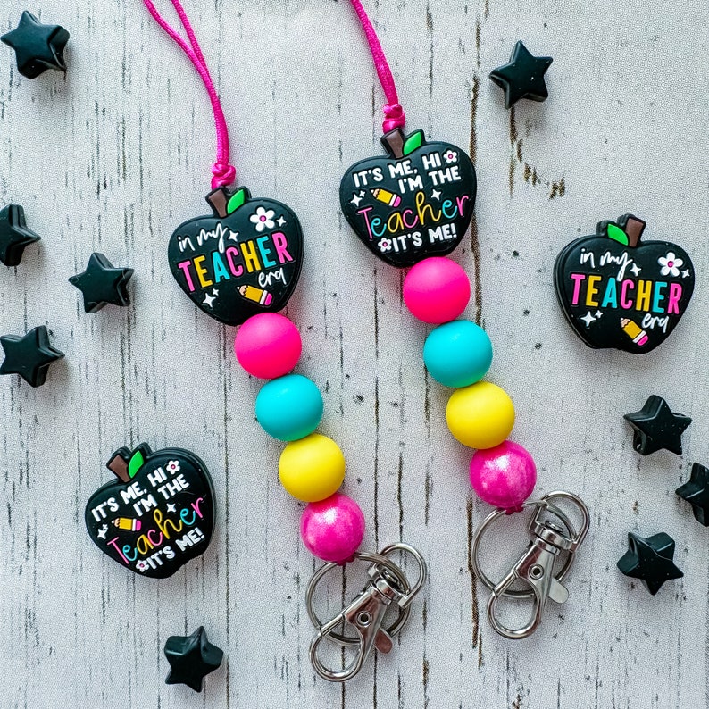 Puede incluir: Dos cordones para profesores con cordones rosas, cuentas de colores y dijes en forma de manzana. Los dijes tienen el texto "IT'S ME, HI I'M THE TEACHER IT'S ME!" y "in my TEACHER era". Adornos negros en forma de estrella est&aacute;n dispersos.