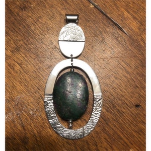 May include: A silver pendant with a green and purple gemstone set in a textured oval frame. The pendant is suspended from a silver bail.