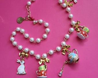 Può includere: Una collana di perle bianche con dettagli dorati e ciondoli. I ciondoli includono una teiera, una tazza da tè e un coniglio. La collana è su uno sfondo rosa.