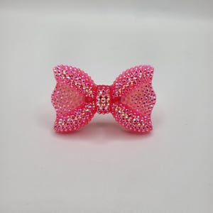 May include: A bright pink bow-shaped hair accessory, covered in sparkling rhinestones. The bow is centered and has a symmetrical design, with a slightly translucent center. The rhinestones catch the light, creating a dazzling effect.