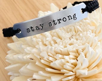 May include: A silver-toned metal bar bracelet featuring the stamped text "stay strong" in black lowercase letters. The metal bar is attached to a black cord band, which is woven with a textured pattern and secured through holes at each end.