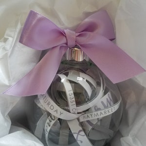May include: A clear glass ornament with a lavender ribbon tied around it. The ornament is filled with white paper strips with text that includes "Saturday", "Haymarket", and "15351 Ch".
