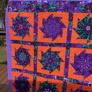 May include: A purple and orange quilt with a geometric pattern. The quilt is made up of squares with a starburst design in the center. The quilt is quilted with a simple stitch.