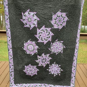 May include: A quilt with a black background and a purple and white floral pattern. The quilt is made up of several different shapes and sizes, and the floral pattern is repeated throughout the quilt.