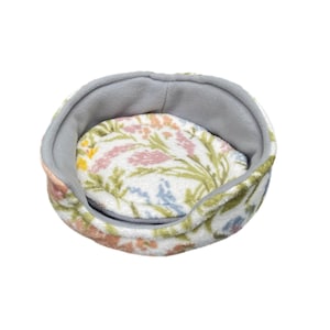 May include: A round pet bed with a floral pattern. The bed has a gray interior and a white exterior with a pattern of pink, yellow, and blue flowers and green leaves. The bed is made of a soft, plush material.