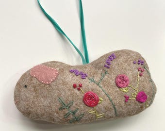 May include: A tan felt bunny ornament with embroidered flowers in pink, red, and purple. The bunny has a pink ear and a teal ribbon for hanging. The floral embroidery includes roses and lavender.