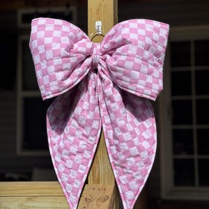 Peut inclure: Un grand nœud en tissu matelassé avec un motif à carreaux rose clair et blanc. Le nœud présente deux larges boucles volumineuses sur le dessus et deux longs pans pointus qui pendent, suspendu par un petit anneau métallique sur un poteau en bois.