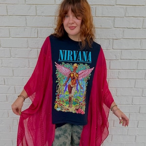 May include: A black short-sleeve t-shirt featuring the word NIRVANA in blue text above a colorful graphic of a winged figure in a forest, layered under a sheer, bright magenta open-front kimono with a textured, patterned fabric finish.