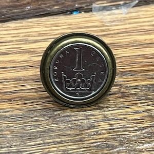 Puede incluir: Un anillo de moneda de color dorado con una moneda checa de 1 Koruna con una corona y el texto "KORUNA CESKA".