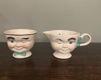 May include: Two white ceramic mugs with a winking face design. The mugs are decorated with a blue bow and the text "Bailey's".