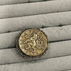 Puede incluir: Una moneda de color dorado con un diseño de león y el texto "CESKA REPUBLIKA" y "2000" alrededor del borde. La moneda está rodeada por un marco metálico decorativo.