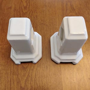 May include: Two white ceramic bookends with a glossy finish. Each bookend features a square-topped pillar design with a stepped, octagonal base and a hollow rectangular cutout on one side, providing a clean and uniform appearance for shelf organization.