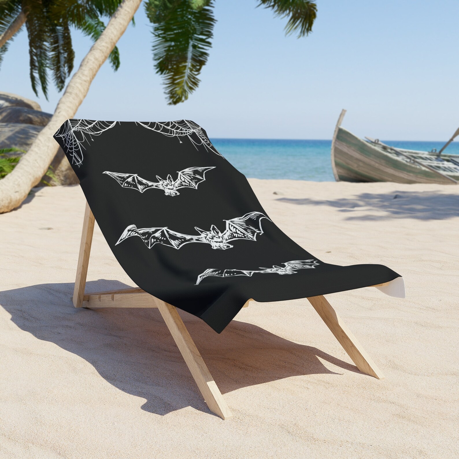 Black beach towel with white bat and spiderweb design draped over a chair on the sand, ideal for summer goth style.