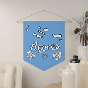 Könnte beinhalten: Ein blaues Banner mit dem Wort "Acores" in weißer Kursivschrift. Das Banner zeigt außerdem eine weiße Umrandung der Azoreninseln, zwei weiße Hortensienblüten und ein weißes Herz mit einem Sonnenstrahl.