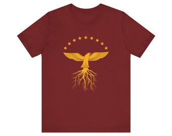 Op de afbeelding: Een maroon t-shirt met een gouden adelaarontwerp. De adelaar is omringd door een cirkel van gouden sterren en heeft wortels die zich uitstrekken vanaf zijn lichaam.