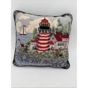 May include: Needlepoint pillow featuring a coastal scene with a red and white striped lighthouse, a sailboat, and a house with a red roof. The pillow has a blue border and depicts people and a dog. The pillow measures approximately 12 inches square.
