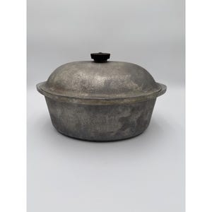 May include: A round, silver-colored metal Dutch oven with a textured, weathered finish. The pot features a matching domed lid with a small, circular black knob centered on top. The vessel has a wide, shallow basin and a flat rim edge.