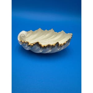 May include: A white porcelain seashell-shaped dish with gold trim. The dish has a wavy, textured design and a curled end. The interior is smooth and white, contrasting with the gold-painted edges. The dish is set against a blue background.
