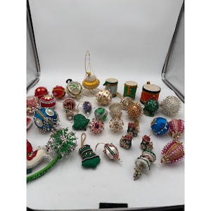 Puede incluir: Una colección de adornos artesanales variados con formas de esferas, campanas y tambores. Estos artículos están decorados con cuentas de colores, lentejuelas, hilos metálicos y texturas de tela en tonos rojo, azul, verde, dorado y blanco.