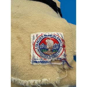 Może przedstawiać: Zbliżenie kremowego koca z czarno-białym paskiem. Na kocu wszyta jest prostokątna etykieta z napisem "Glacier Park Blanket" oraz sceną g&oacute;rską z owcą.