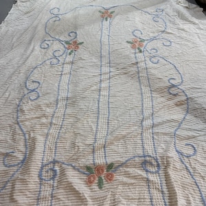 May include: Cream-coloured fabric with a textured, striped pattern. The design features light blue embroidered lines and clusters of pink roses. The embroidered elements create a decorative, floral pattern across the fabric.